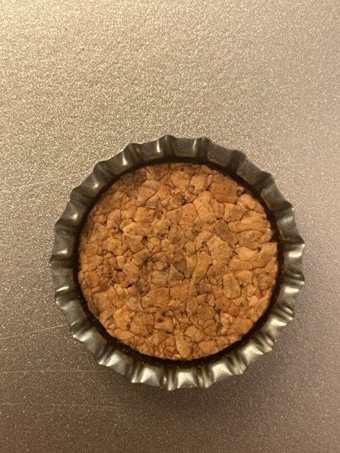 Overhead view of an antique cork-lined bottle cap with ridged metal crown edges and tan cork interior, on a beige textured surface.