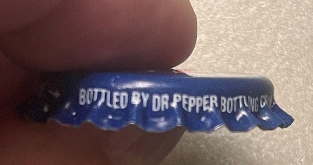 Close-up of blue bottle cap held by thumb, white text reads "BOTTLED BY DR PEPPER BOTTLING CO" with partial location visible