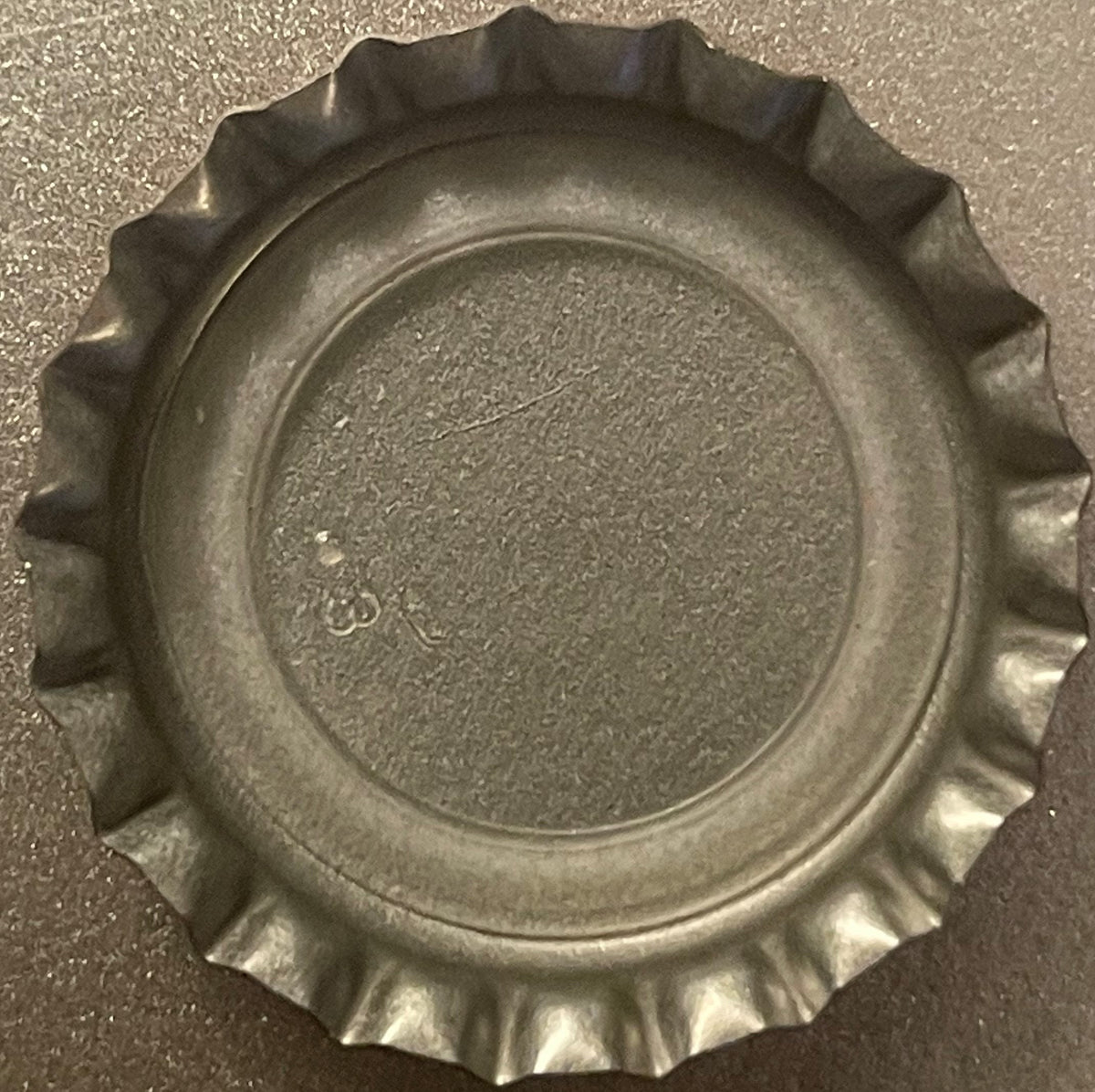 Metallic bottle cap with crimped edges viewed from above on textured gray surface, embossed circular center detail visible