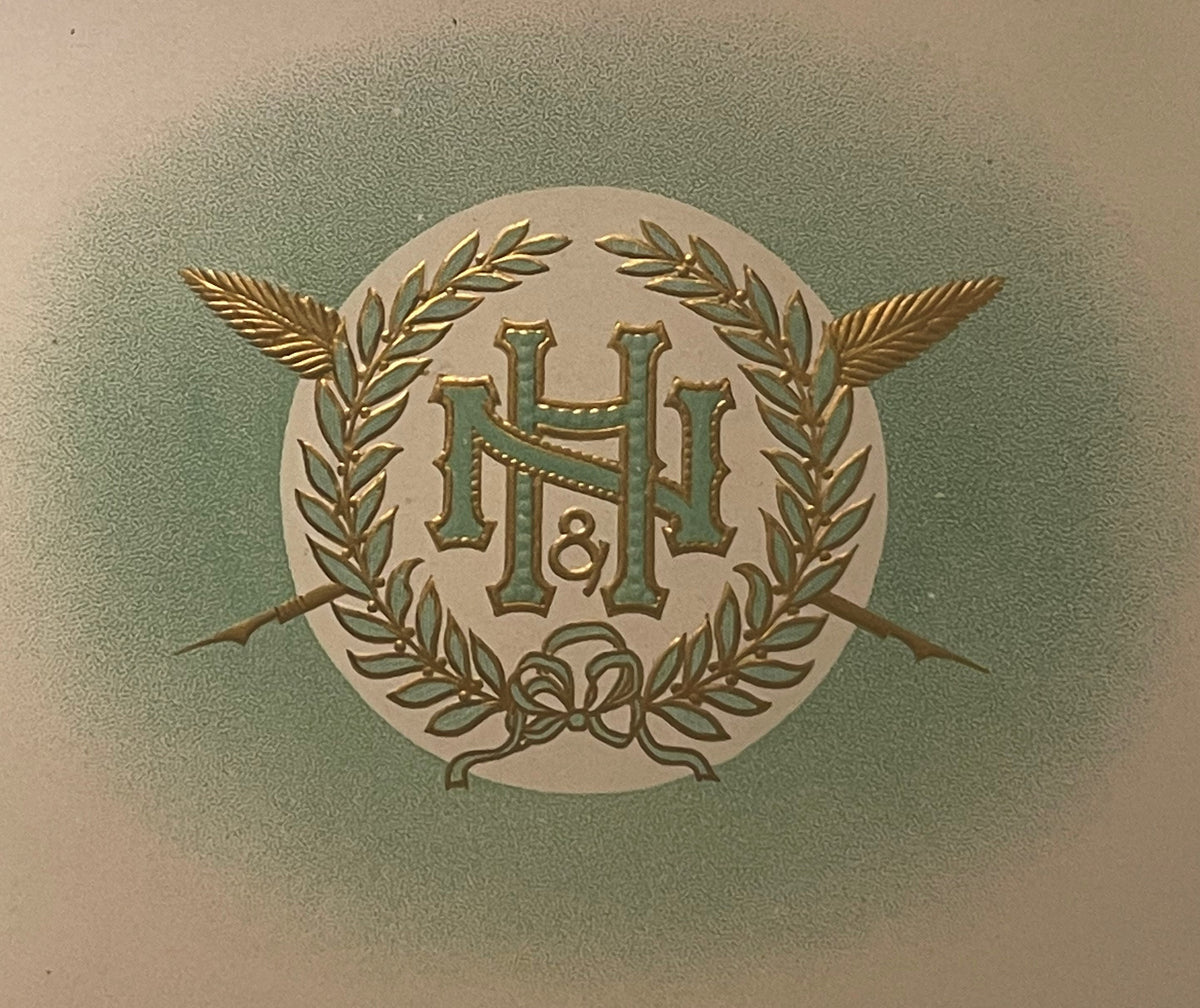 Close-up of the N&H cigar label: an interlaced green and gold N and H monogram with a small ampersand, inside a gold laurel wreath tied with a bow, with two crossed feathered gold shafts, on a pale circle in a green haze