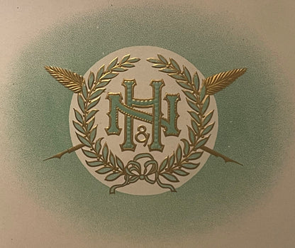 Close-up of the N&H cigar label: an interlaced green and gold N and H monogram with a small ampersand, inside a gold laurel wreath tied with a bow, with two crossed feathered gold shafts, on a pale circle in a green haze