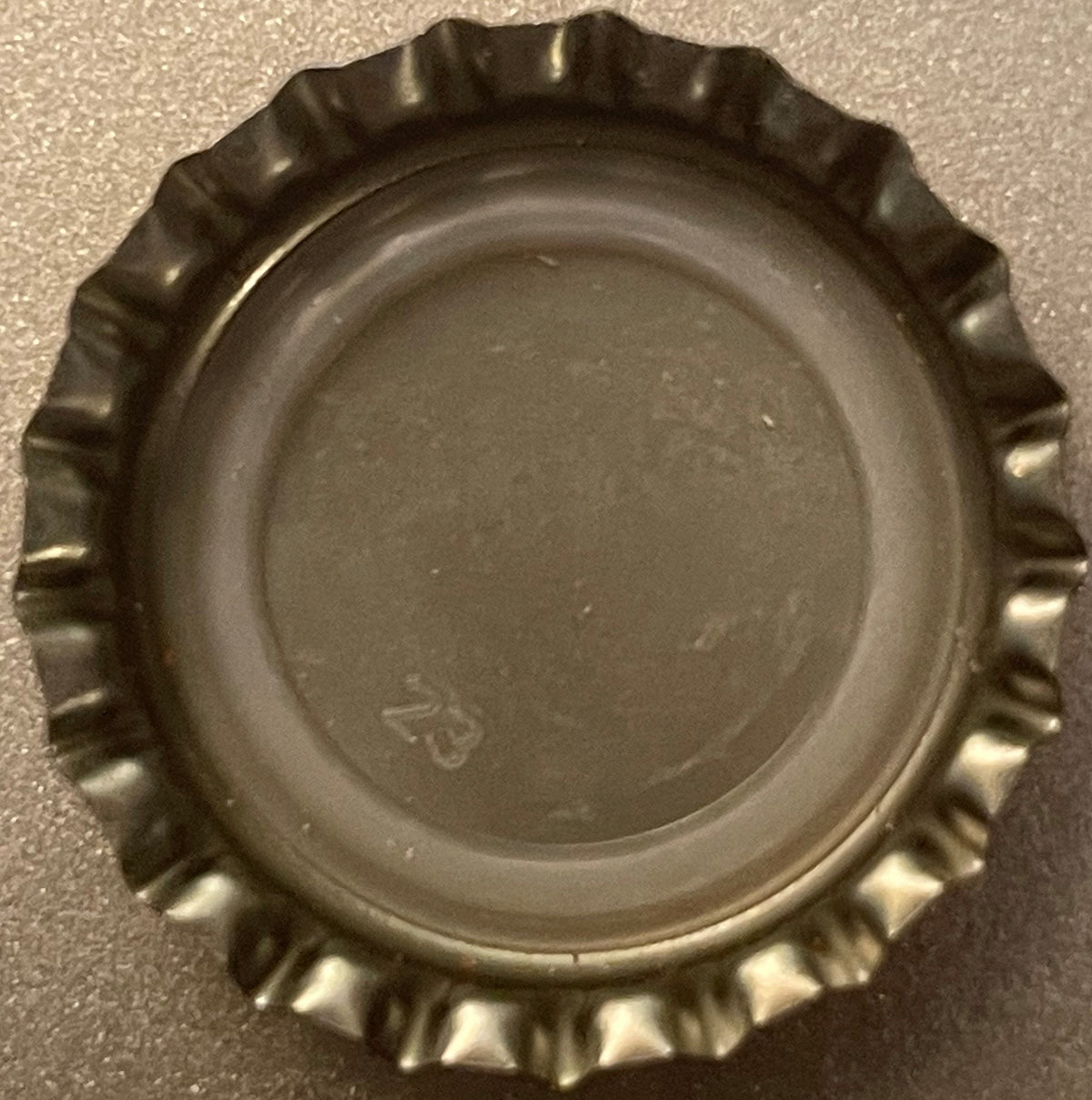 Vintage metal bottle cap with crimped edges, shown from above on gray surface, displaying embossed logo in center
