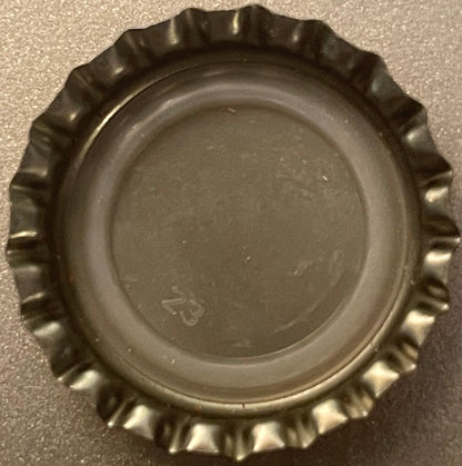 Vintage metal bottle cap with crimped edges, shown from above on gray surface, displaying embossed logo in center