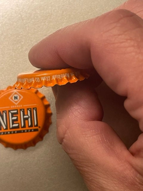 Close-up of orange Nehi bottle cap held between two adult fingers, showing embossed text and bottling company mark from West 