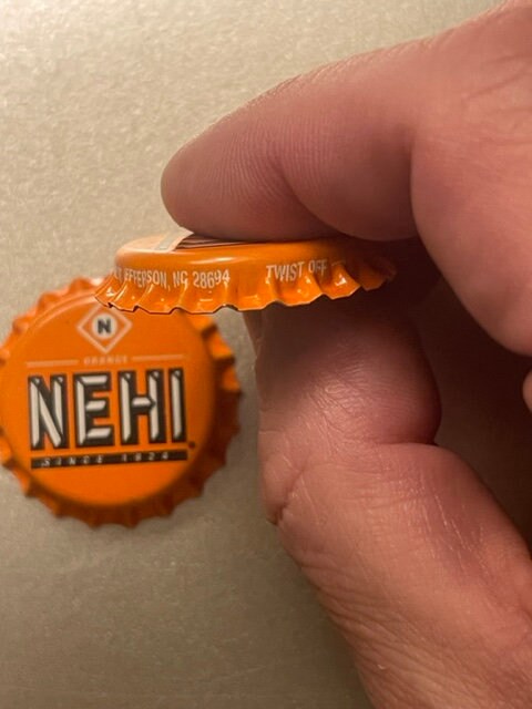 Orange Nehi bottle cap held in fingers showing embossed text and maker's mark, with partial address visible on crimped edge