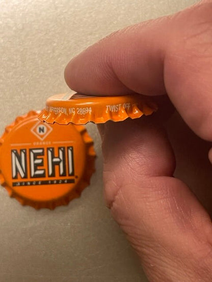 Orange Nehi bottle cap held in fingers showing embossed text and maker's mark, with partial address visible on crimped edge