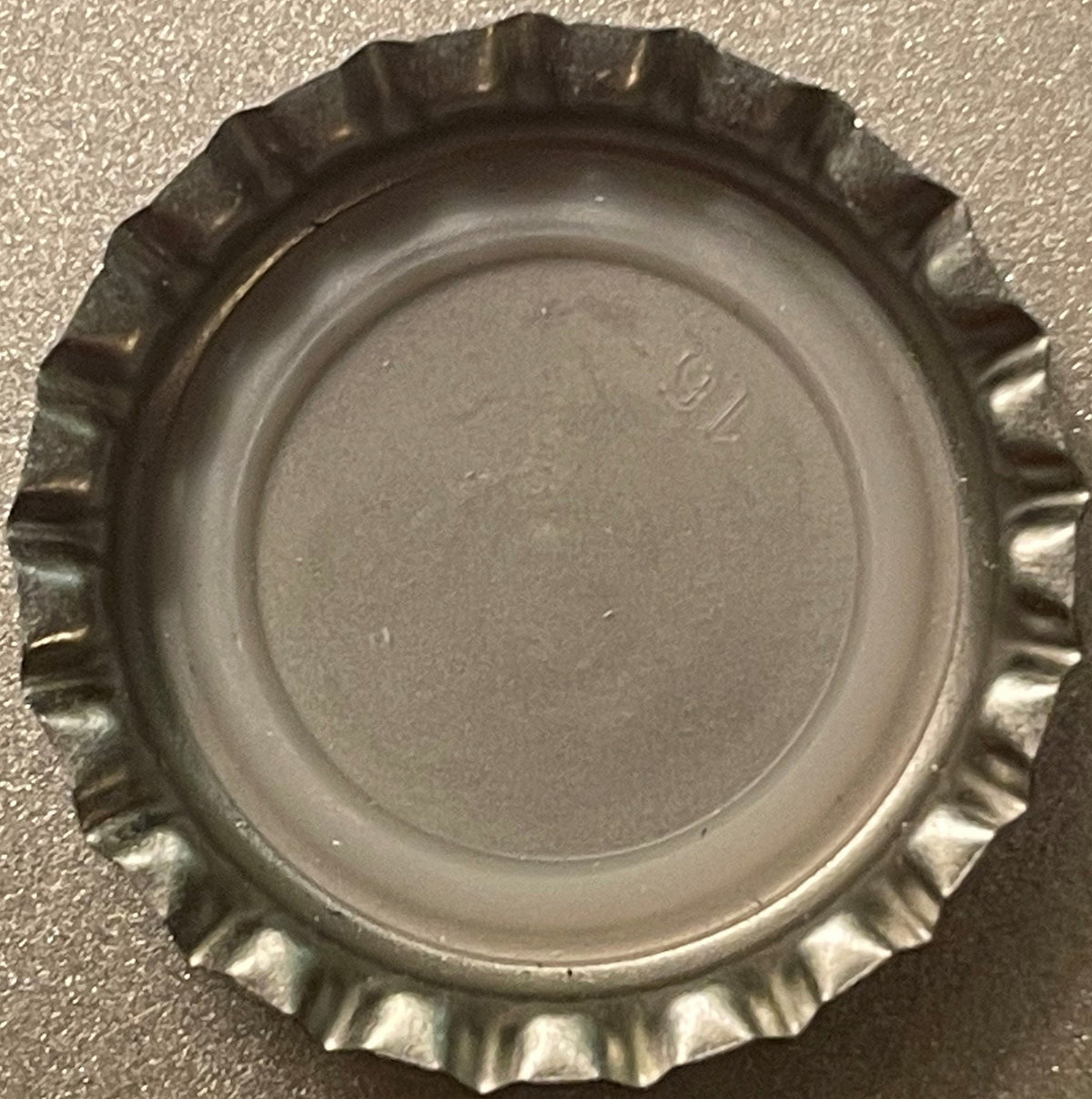 Vintage metal bottle cap with crimped edges and embossed lettering, photographed flat against gray textured surface