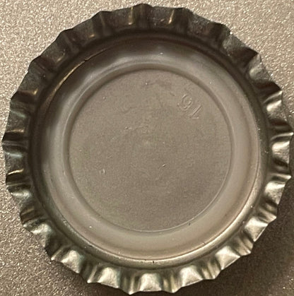 Vintage metal bottle cap with crimped edges and embossed lettering, photographed flat against gray textured surface