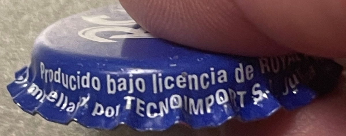 Close-up of blue RC Cola bottle cap held by a thumb, showing embossed text "Producido bajo licencia de" and "TECNOIMPORT" par