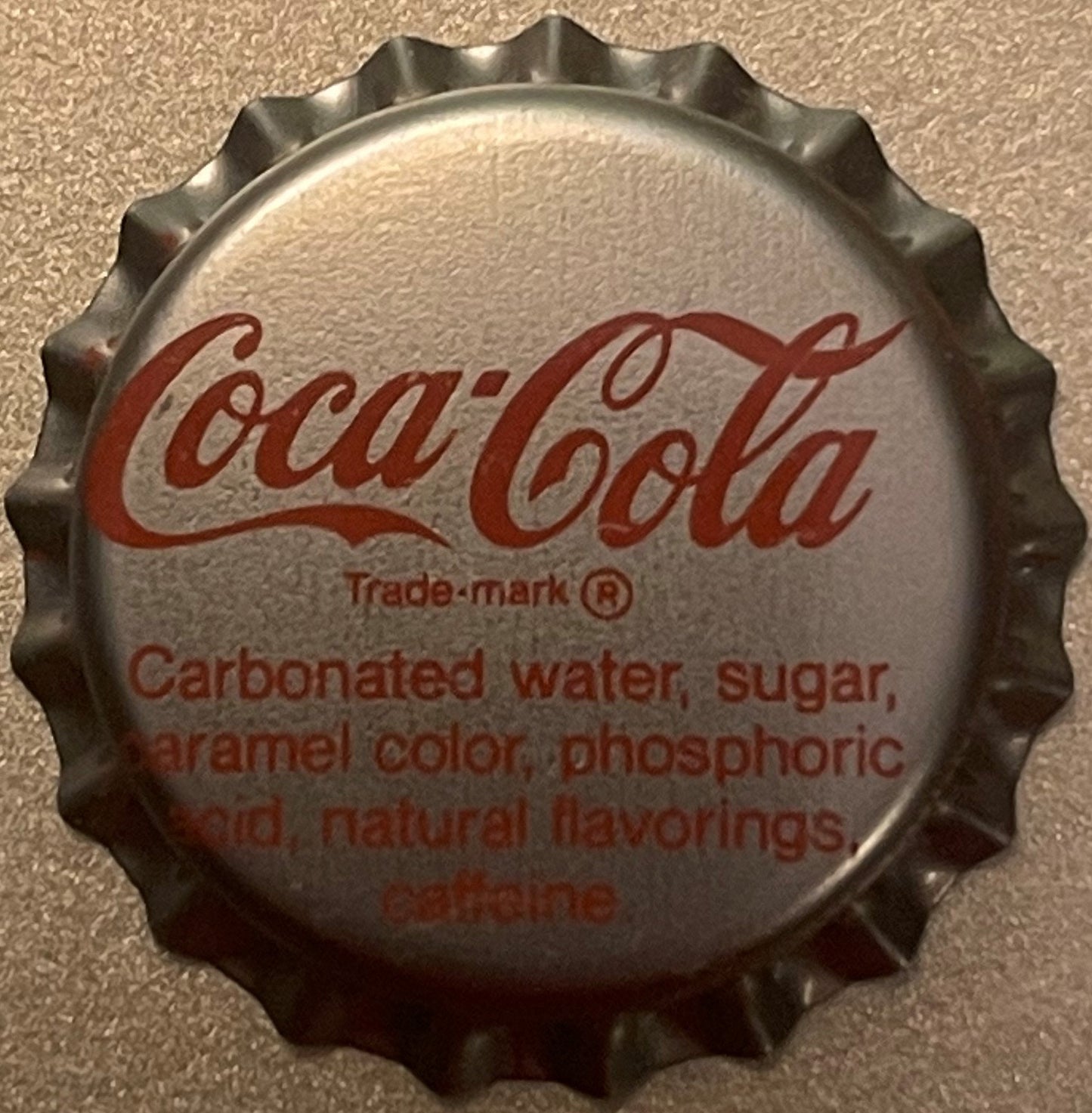 Silver Coca-Cola bottle cap with red script logo and ingredients list, photographed flat against beige textured surface