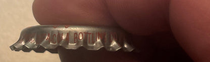 Close-up of a vintage metal bottle cap held by a thumb, showing embossed text including "BOTTL" and partial ingredients list 