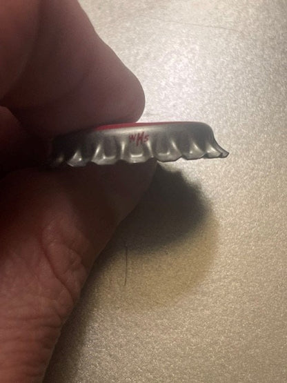 Edge view of the unused Cherry Soda crown cap held in the fingers: the fluted metal skirt with the cap maker's small red WHS mark