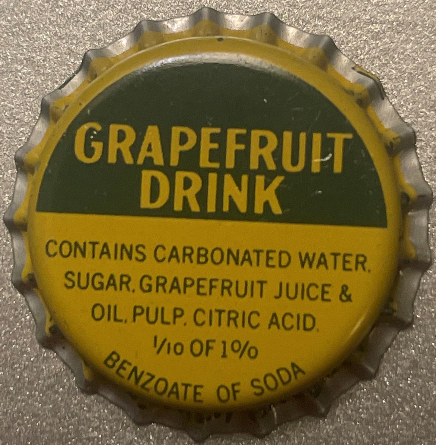Vintage yellow and green grapefruit drink bottle cap with embossed crown edge and printed text on concrete surface.