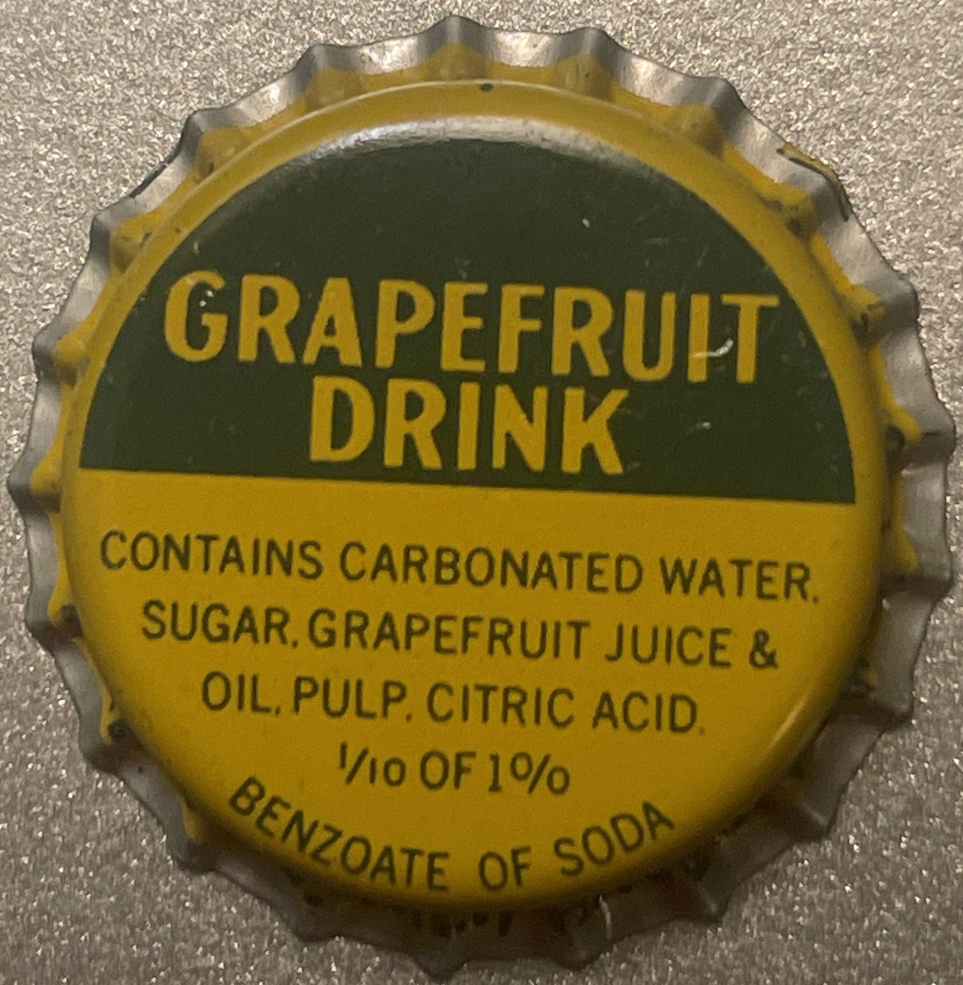 Vintage yellow and green grapefruit drink bottle cap with embossed crown edge and printed text on concrete surface.