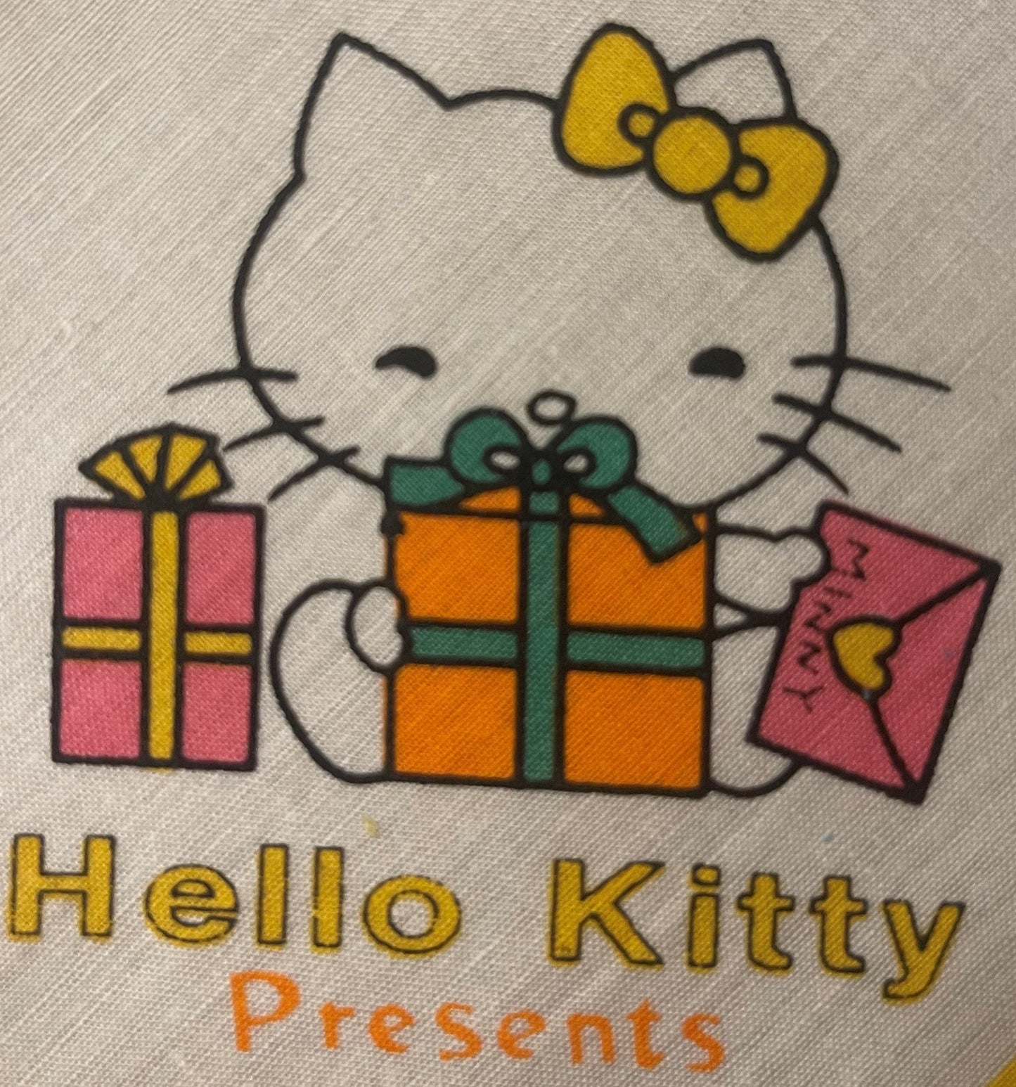 Hello Kitty with yellow bow holding three wrapped presents, printed on cream cotton fabric with yellow text below reading "He