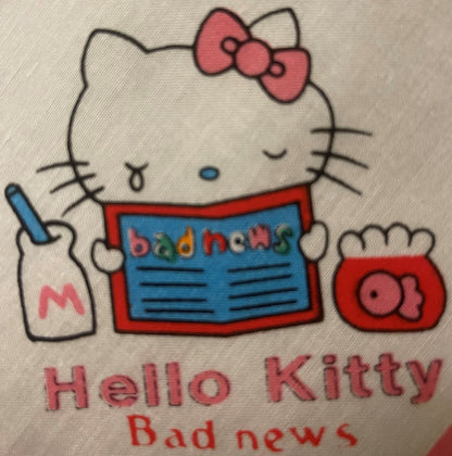 Hello Kitty cotton napkin with pink bow, holding newspaper labeled "bad news," red jar beside her, printed text reads "Hello 