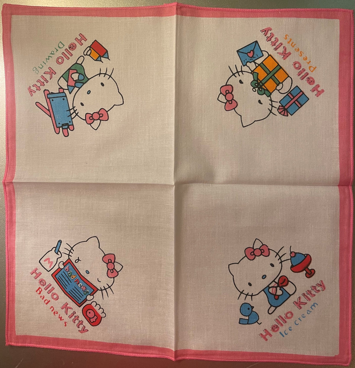 Unfolded pink-bordered cotton napkin displaying four Hello Kitty illustrations with colored text on cream fabric, laid flat o