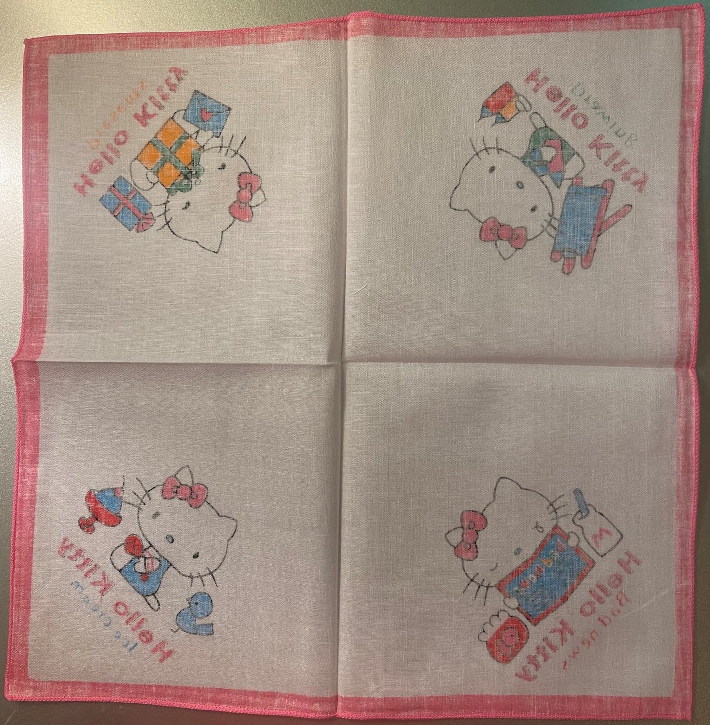 Unfolded pink-bordered cotton napkin with four Hello Kitty illustrations in different poses holding gifts and presents, print