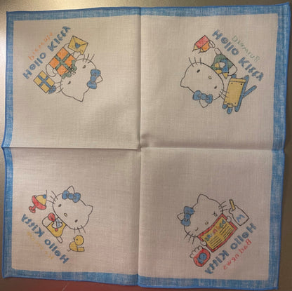 Unfolded cotton napkin with blue border showing four Hello Kitty illustrations in corners with colored kites, toys, and Japan