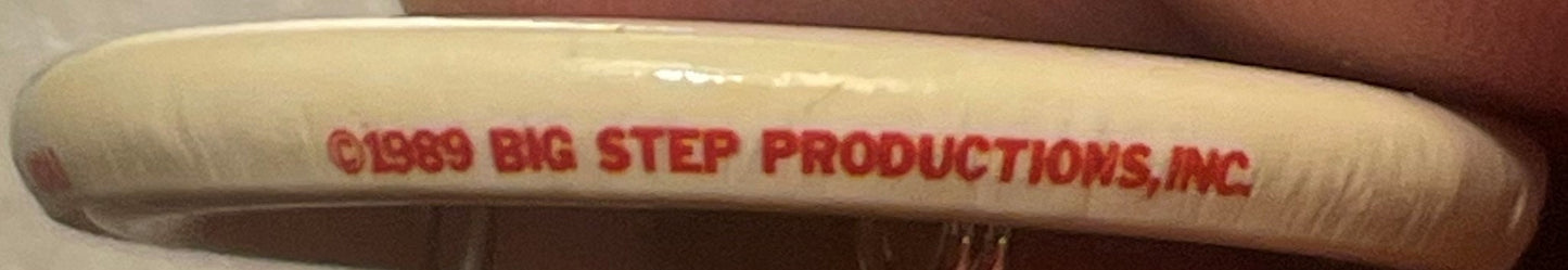 Close-up of vintage pin showing red text reading "©1989 BIG STEP PRODUCTIONS, INC" on cream-colored cylindrical surface