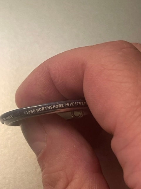 Close-up of vintage pin held between fingers showing embossed text "1990 NORTHSHORE INVESTMENT" on dark metal backing