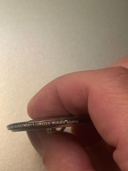 Close-up of vintage TMNT pinback button held between thumb and finger, showing embossed text reading "INVESTMENTS LIMITED MIR