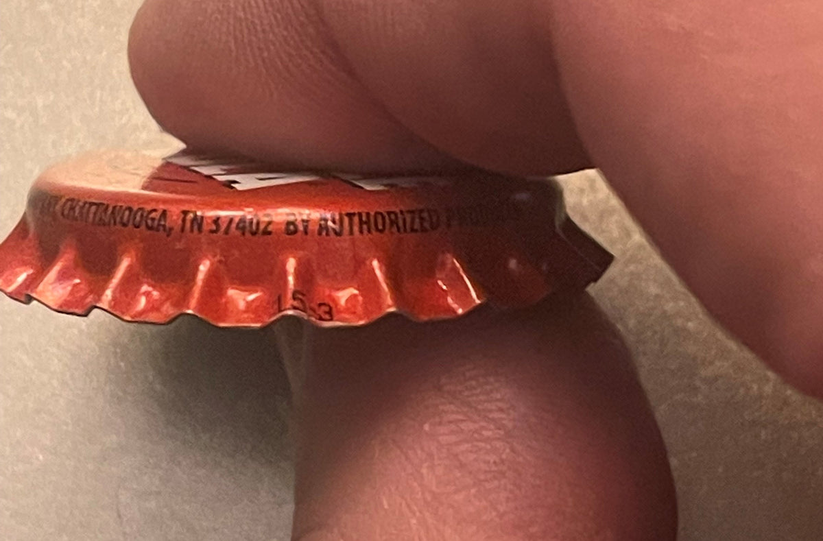 Red bottle cap held by a thumb showing embossed text reading "CHATTANOOGA, TN" and "AUTHORIZED," close-up detail view