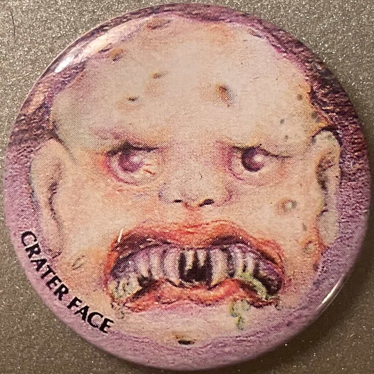 Circular pinback button with grotesque face featuring prominent teeth, deep facial craters, and reddish-brown beard on pale s
