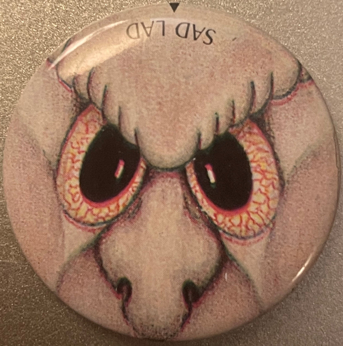 Circular pin-back button featuring a pale droopy face with large bloodshot eyes outlined in pink and yellow, black pupils, an