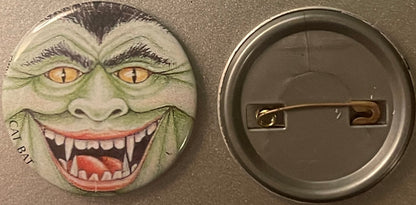 Vintage pinback button with grotesque green vampire face with fanged grin and yellow cat eyes, shown front and back against g
