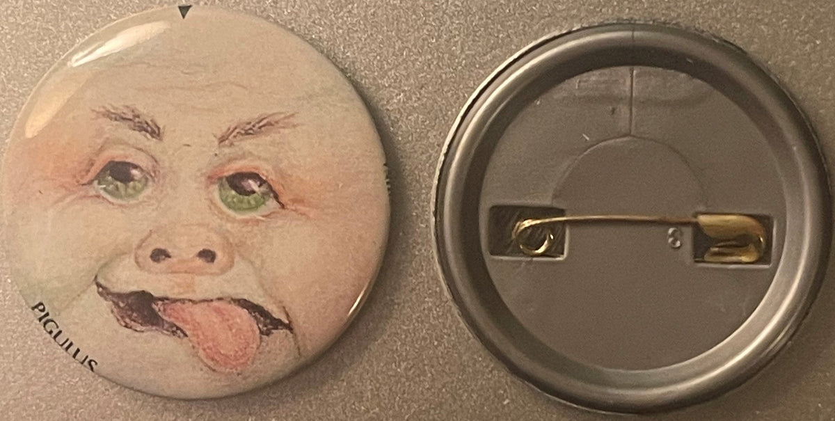 Front and back of a round pinback button featuring a grotesque baby face with green eyes, protruding tongue, and "PIGULUS" te