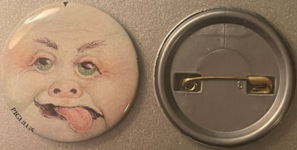 Front and back of a round pinback button featuring a grotesque baby face with green eyes, protruding tongue, and "PIGULUS" te