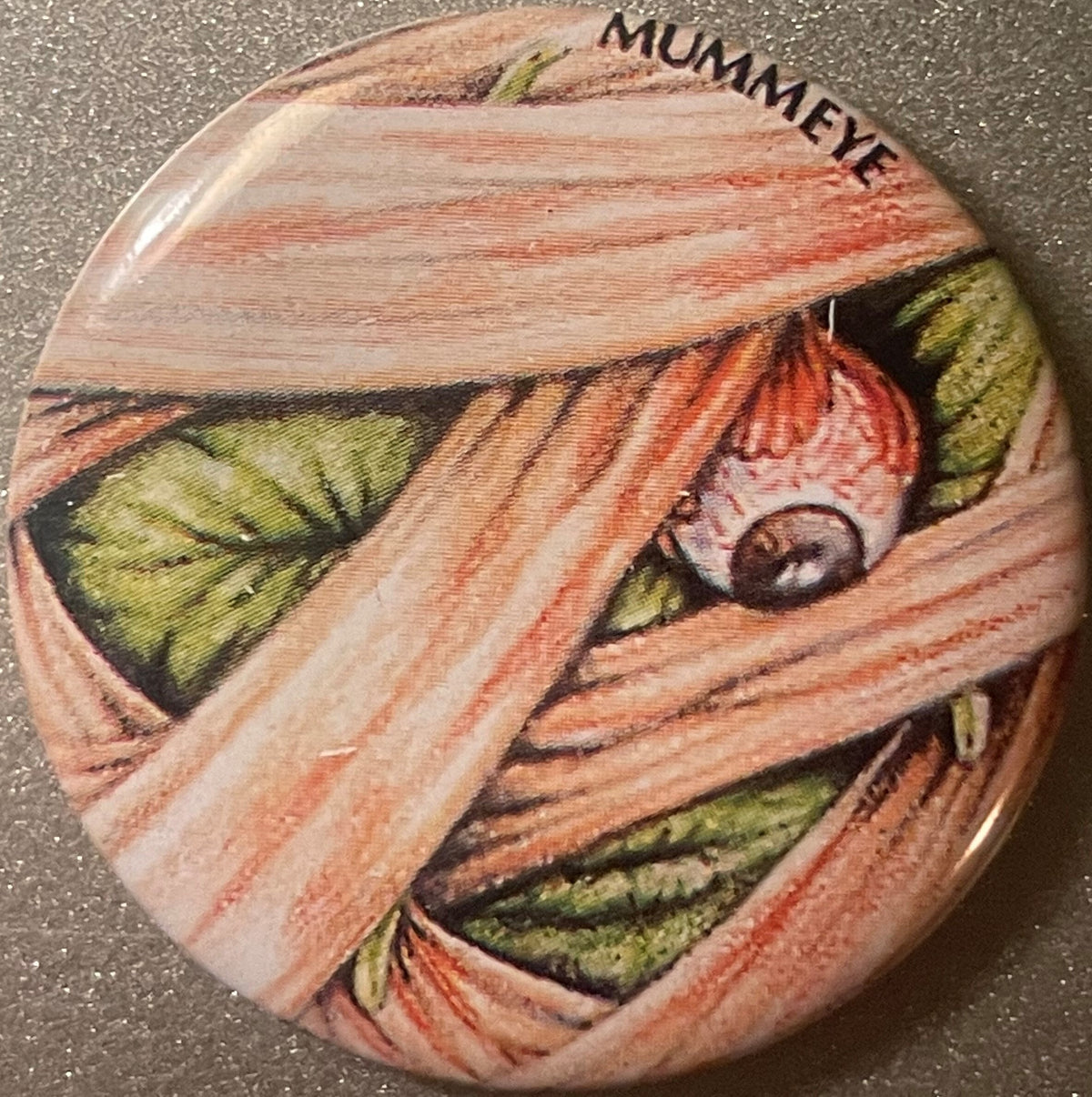 Round vintage pin with mummy-wrapped face design in tan, red, and green, with dark eye visible and "MUMMEYE" text at top