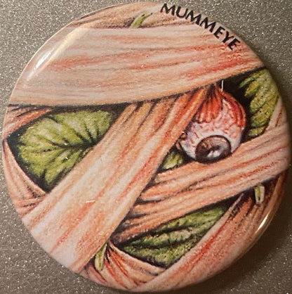 Round vintage pin with mummy-wrapped face design in tan, red, and green, with dark eye visible and "MUMMEYE" text at top