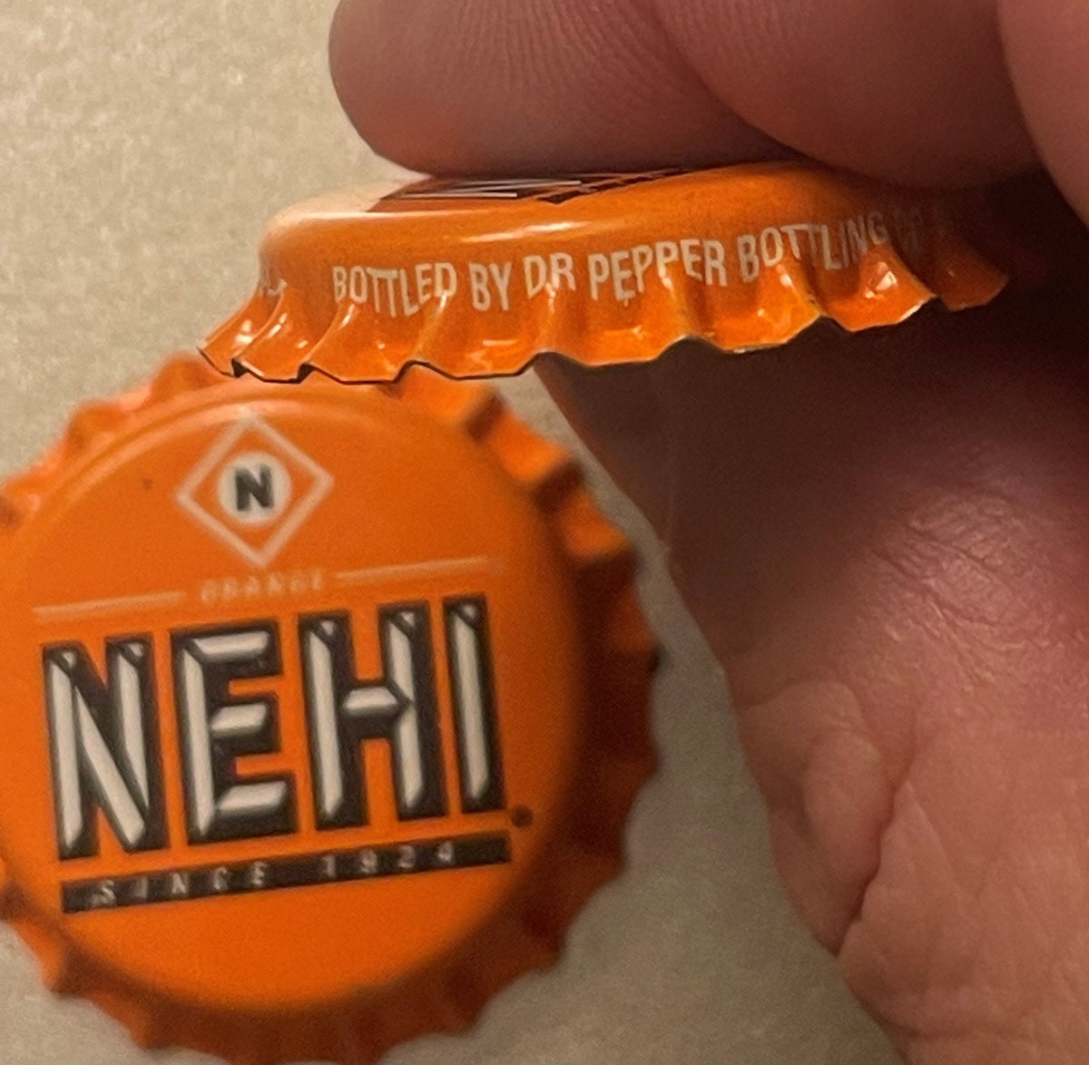 Close-up of orange Nehi bottle cap held in hand, showing "NEHI" text and "Bottled by Dr Pepper Bottling" on crown, with diamo