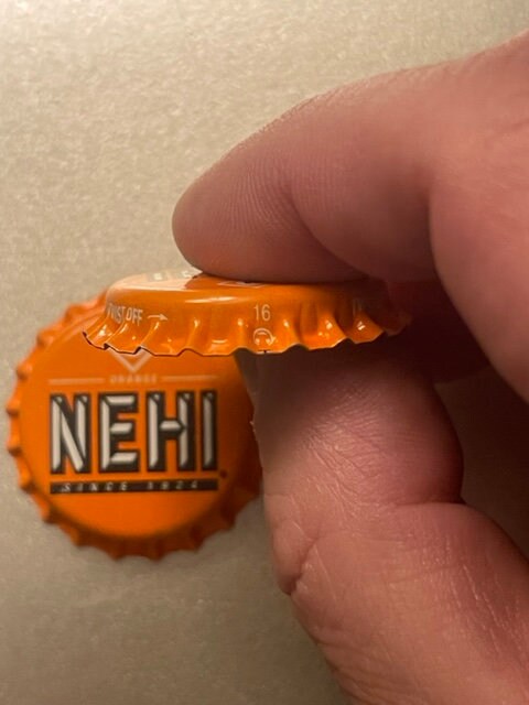 Orange Nehi bottle cap held between thumb and fingers, showing embossed text and ridged edges against beige background