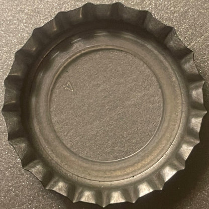 Metallic bottle cap with fluted edges shown flat against textured gray surface, small "A" mark visible on interior