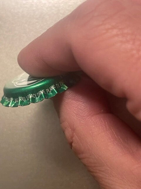 Green twist-off bottle cap held between thumb and fingers for scale, showing embossed text and metallic interior lining