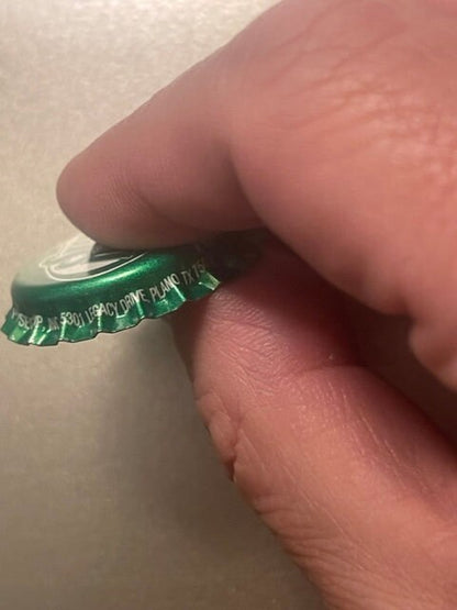 Green twist-off bottle cap held between thumb and fingers for scale, showing embossed text and metallic interior lining