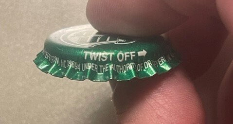 Green twist-off bottle cap held by a finger for scale, showing "Sun Drop" branding and West Jefferson NC text embossed on whi