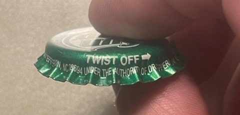 Green twist-off bottle cap held by a finger, showing "TWIST OFF" text and white ridged crown edge against skin for scale