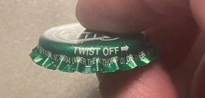 Green twist-off bottle cap held by a finger, showing "TWIST OFF" text and white ridged crown edge against skin for scale