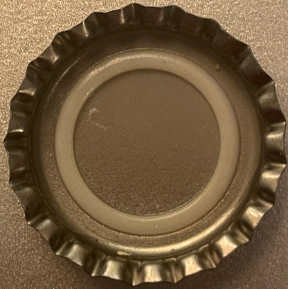 Silver metal bottle cap with ridged edges photographed from above against a textured gray surface, showing smooth center disc