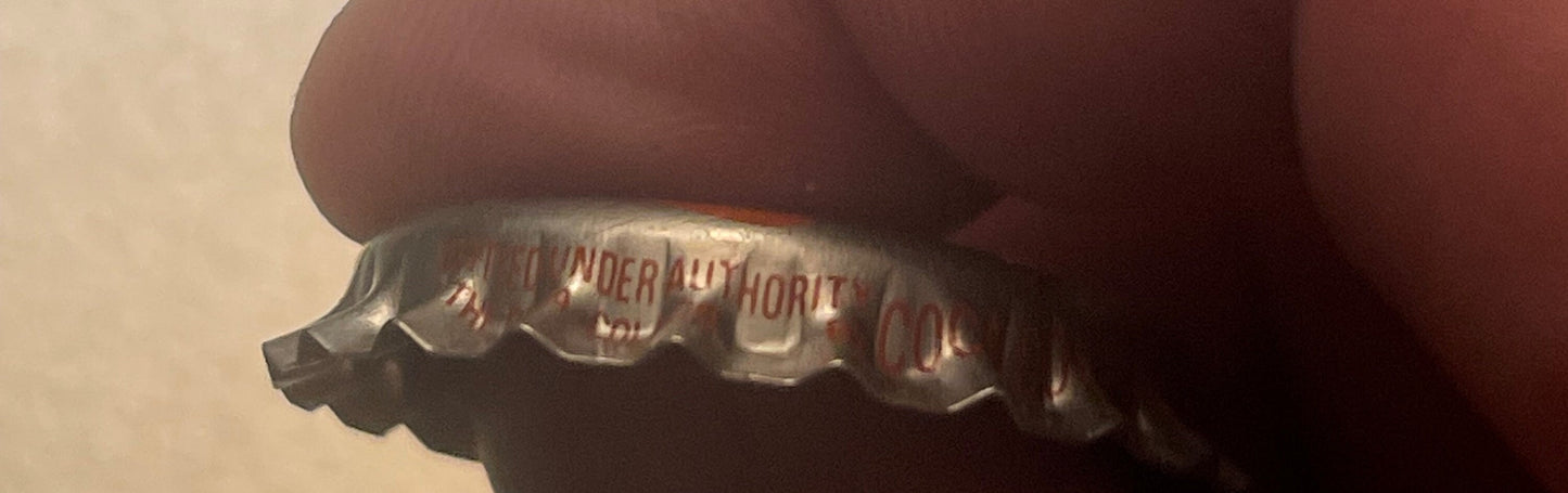 Close-up of a vintage metal bottle cap with embossed text and ingredients list, held against a brown fabric background for sc