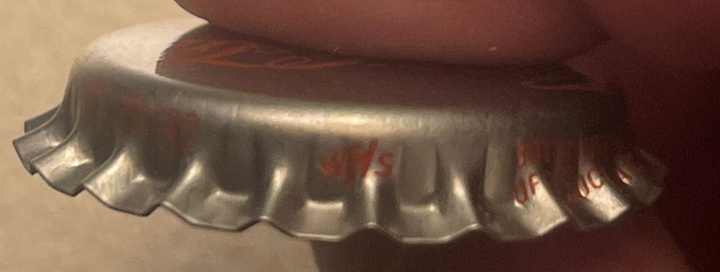 Close-up of a silver lithographed bottle cap with fluted edges and red text on the rim, photographed at an angle against a ta