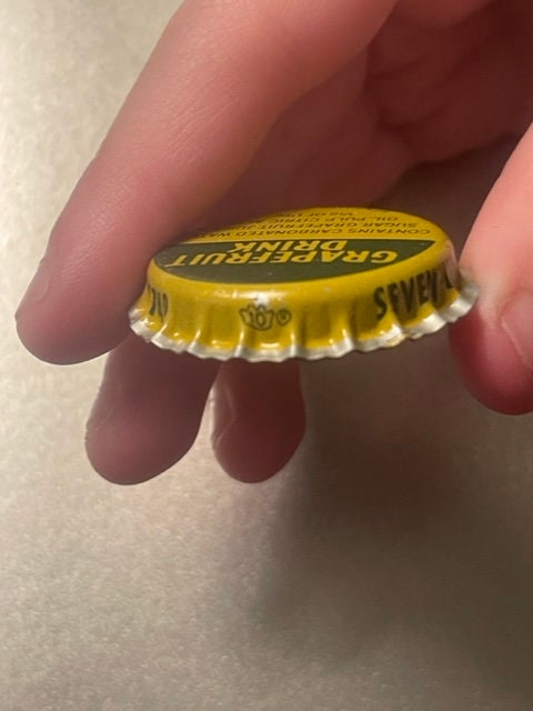 Yellow Seven-Up bottle cap held in fingertips for scale, showing lithographed text and Hagerstown MD marking on crown.
