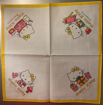 Unfolded yellow-bordered cotton napkin with four Hello Kitty panels showing the character with gifts, makeup items, and text 