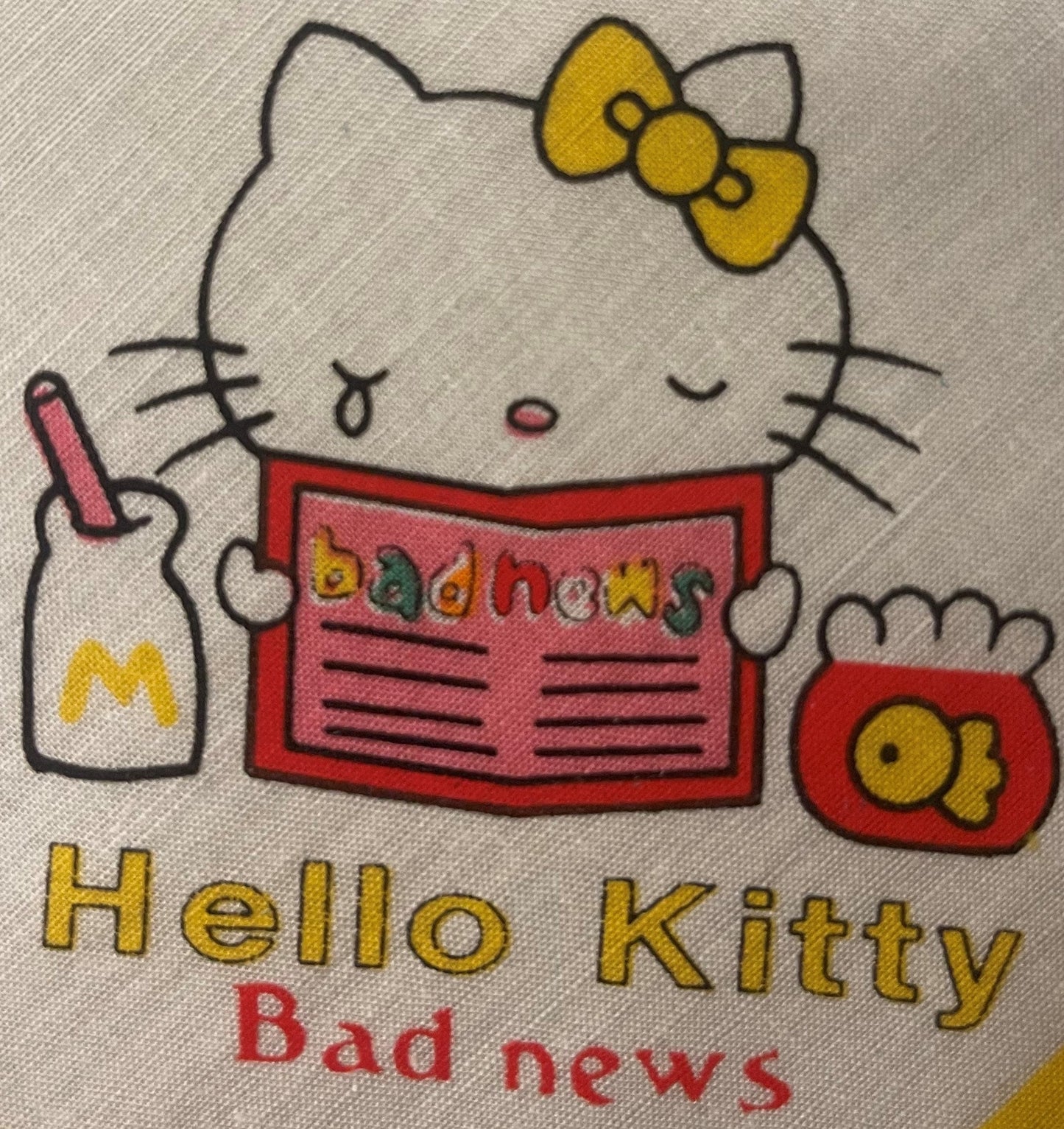 Hello Kitty cotton napkin with yellow bow, holding red newspaper labeled "bad news," against beige fabric background.