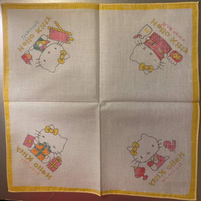 Unfolded cotton napkin with four Hello Kitty panels featuring gifts and bows, yellow border, printed text in multiple languag