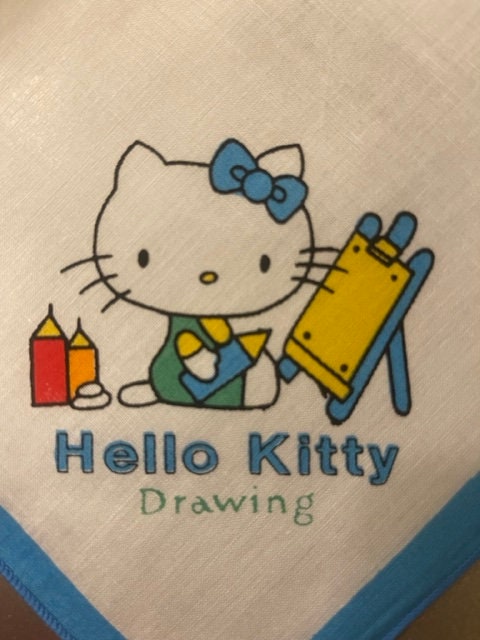 Cream cotton napkin with Hello Kitty holding art supplies, blue bow and text, laid flat on blue fabric edge visible